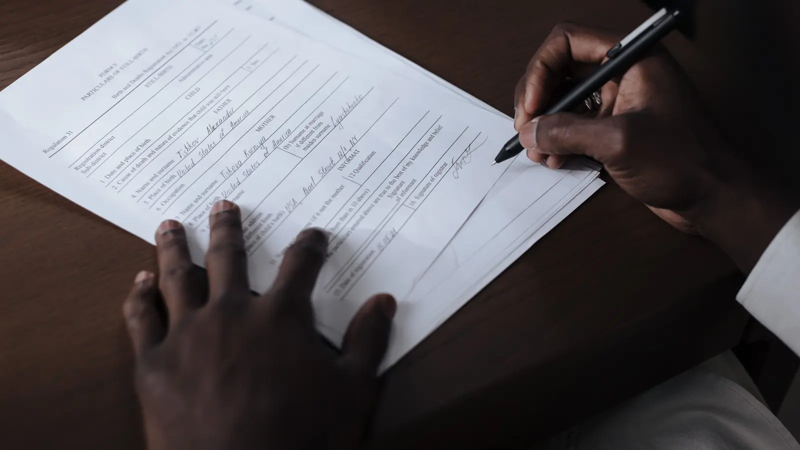 A close-up of hands filling in an official printed form with a pen at a dark wooden desk, one hand steadying the page.