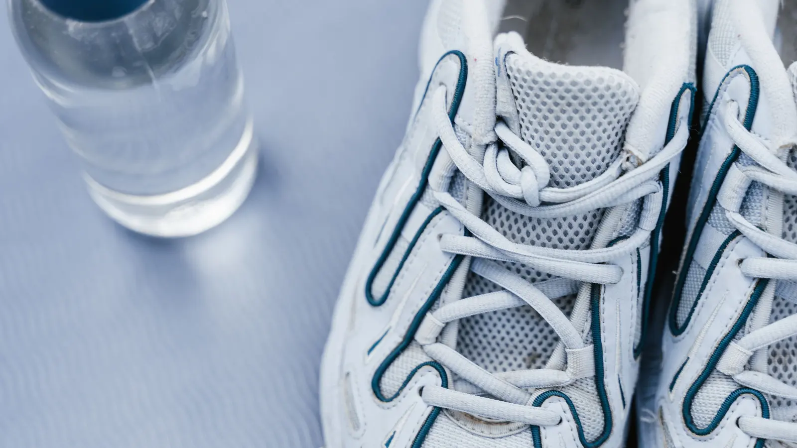 A close-up of white trainers with the laces done up, beside a clear water bottle on a pale surface.