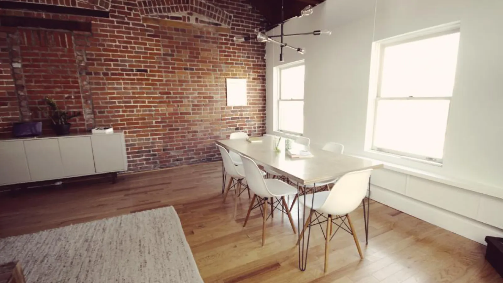 An empty meeting room with an exposed brick wall, a long white table, white moulded chairs, and bright windows along one side.