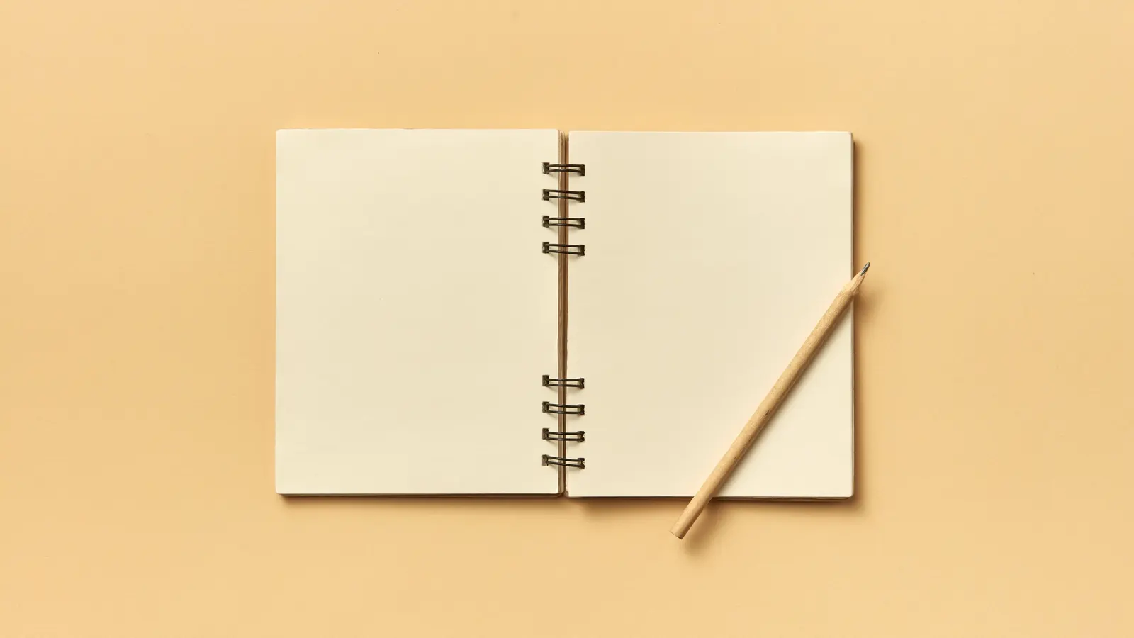 An open spiral-bound notebook with blank cream pages on a warm sand-coloured background, a sharpened wooden pencil resting diagonally across the right-hand page.