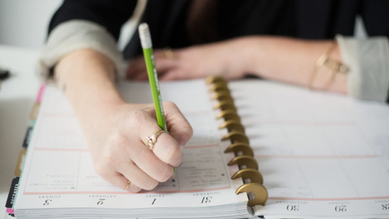 A close-up of a hand writing into a spiral-bound dated planner with a green pencil.