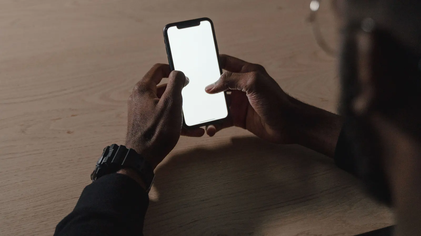 Two hands holding a smartphone with a blank white screen above a wooden desk.