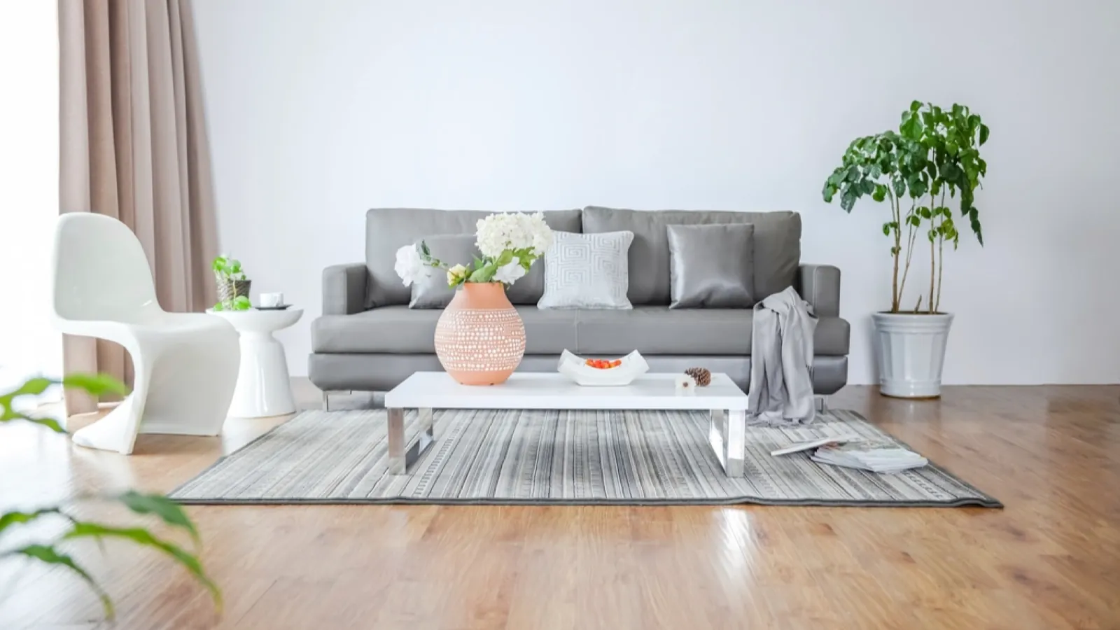A pale, bright living room with a grey sofa, a low coffee table holding a vase, houseplants, and a striped rug on a wooden floor.