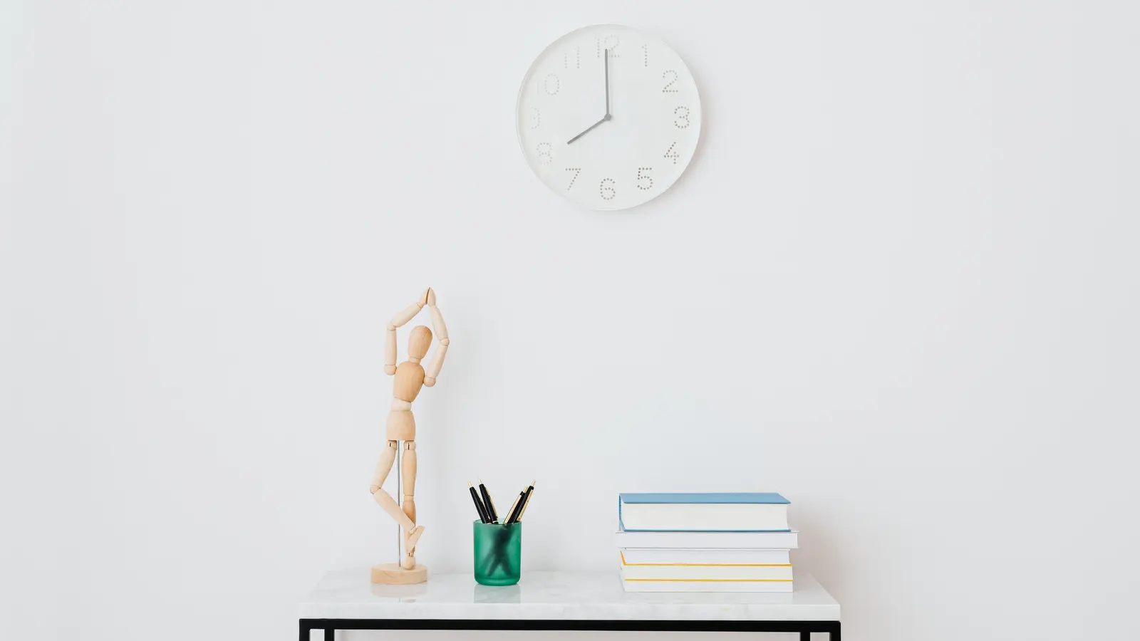 A plain white wall clock above a narrow console table holding a wooden artist's figure, a pot of pens and a stack of books