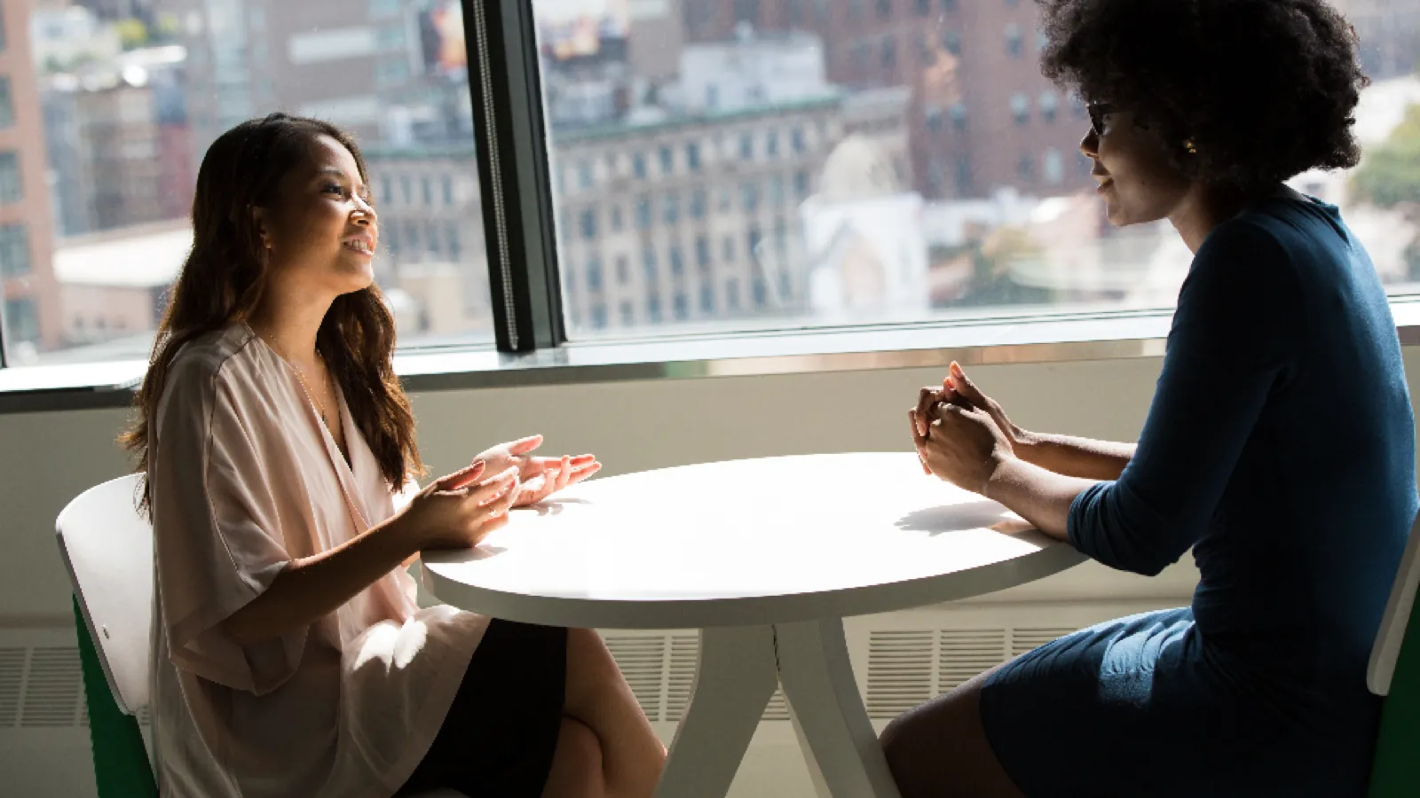 Two women sitting opposite each other at a small round table beside a bright window, talking.