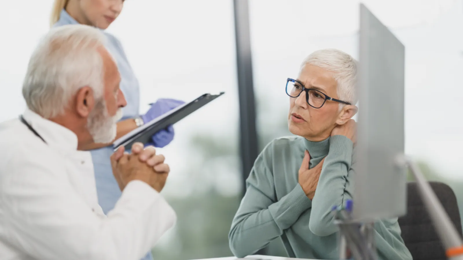 A doctor in a white coat holding a clipboard, listening to an older woman in glasses who is describing her symptoms.
